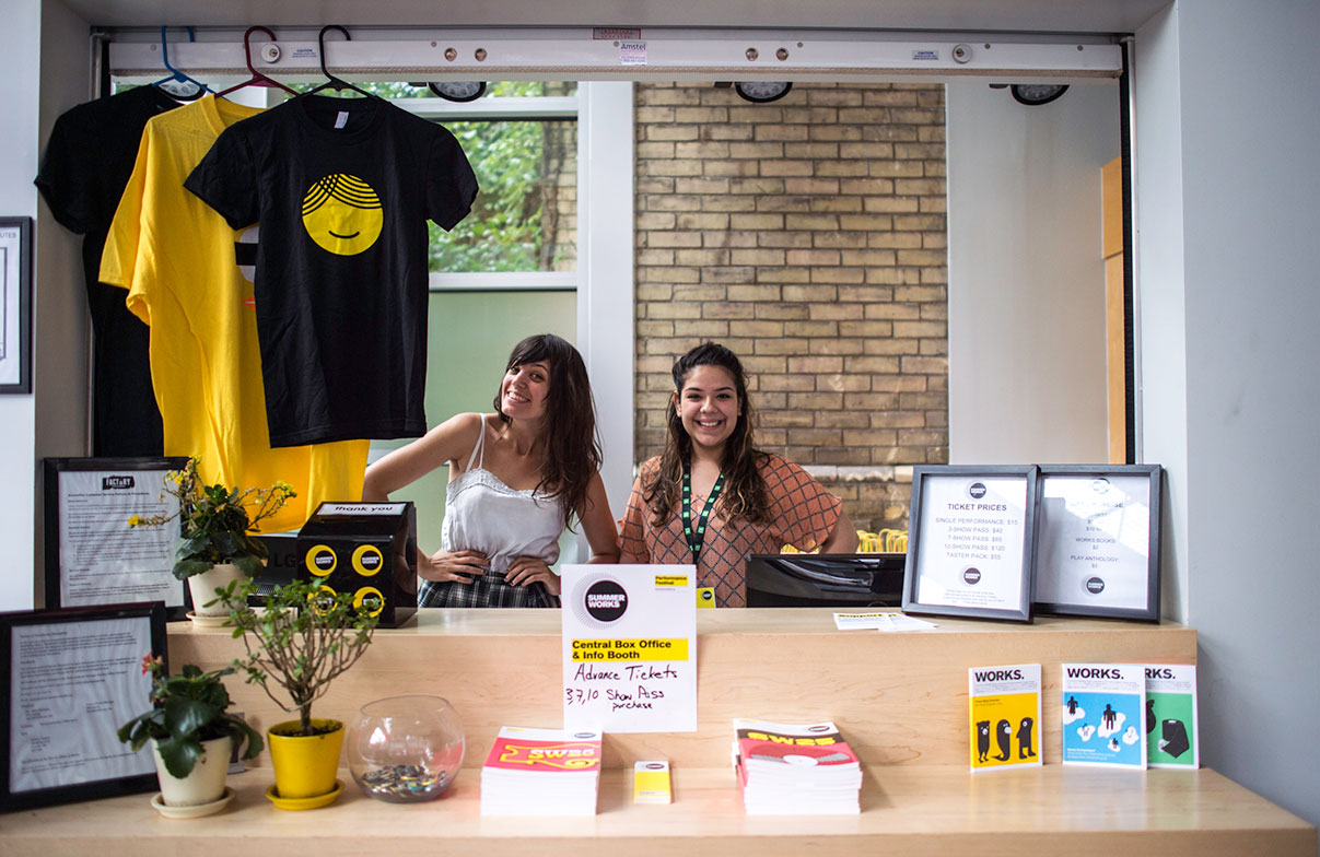 Two young volunteers stand behind a counter full of SummerWorks books and merchandise