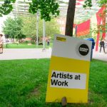 A sign posted in a city park that reads Artists At Work
