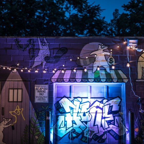 A courtyard at night decorated with strings of lights. A graffiti mural of houses and raccoons is in the background
