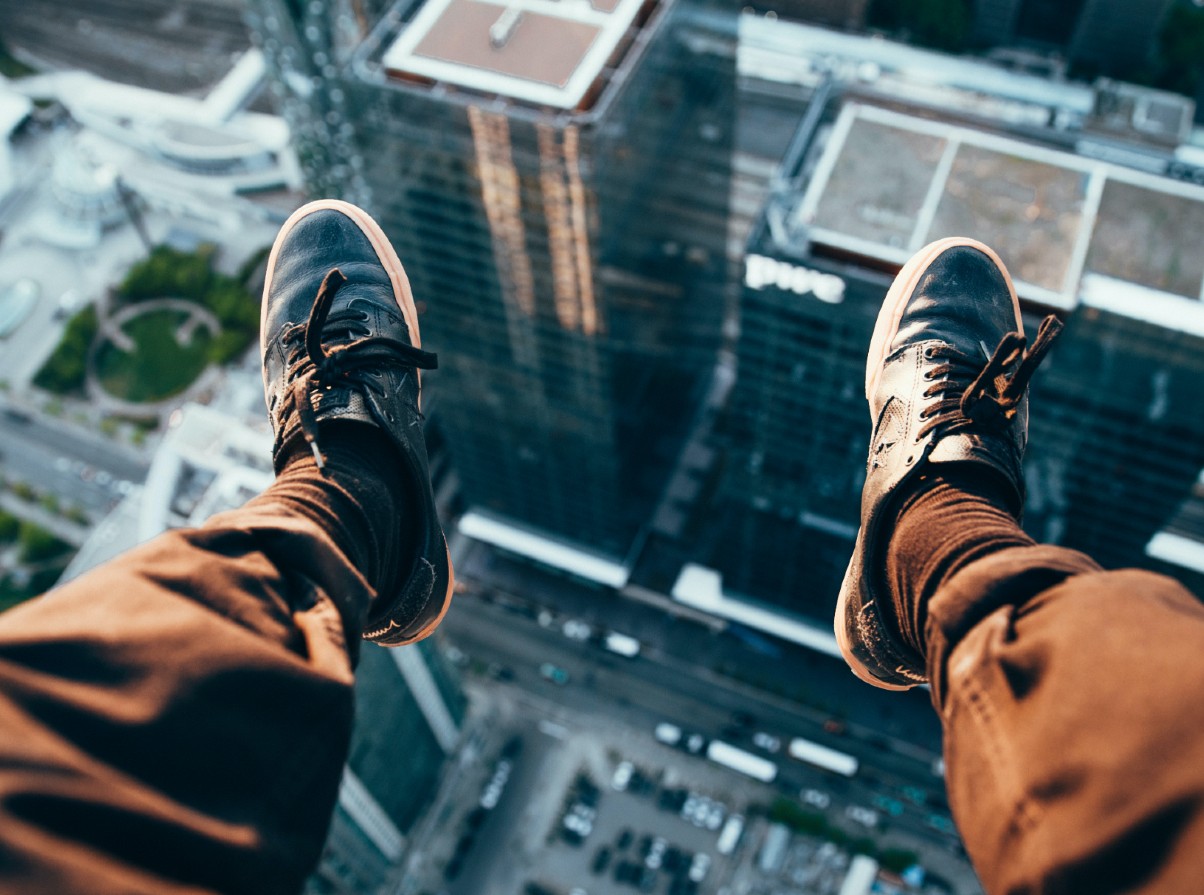 Looking down onto a city street from a great height, a pair of legs in brown pants and black shoes are in the foreground