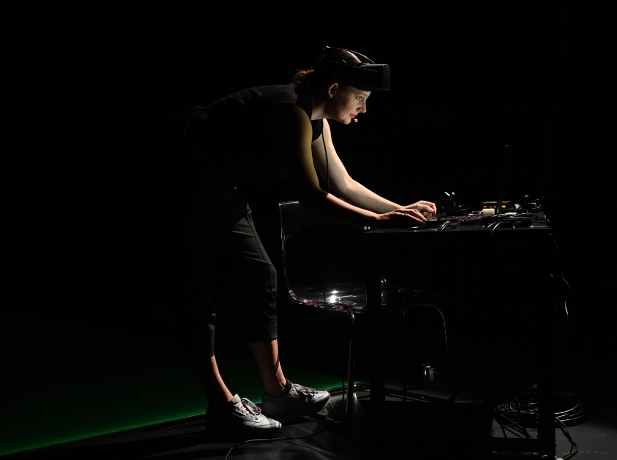 Freya is photographed on stage, bending over a desk looking into a laptop. There is a black table with wires coming down behind the desk and a black chair shining from a spotlight on top. Freya has a AR helmet while wearing all black and grey runners. The floor is black with a green screen showing behind.