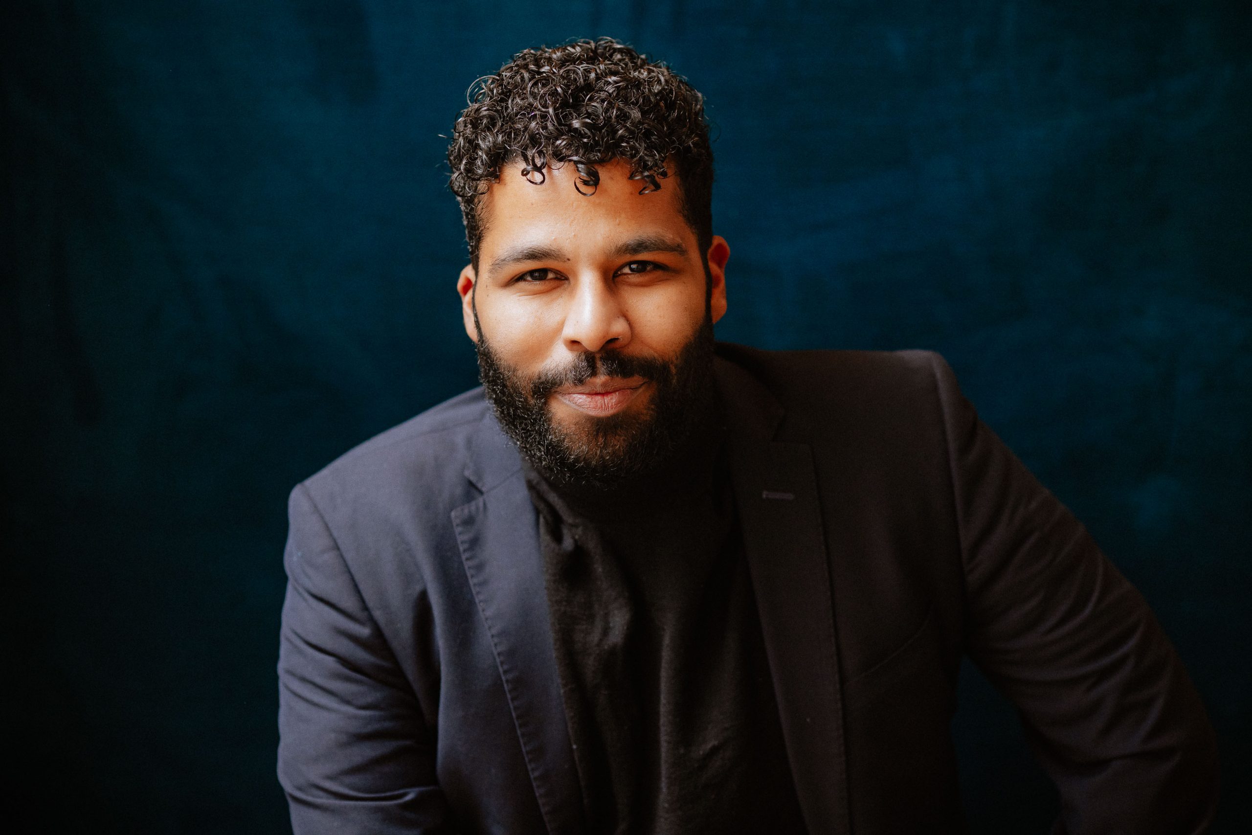 Jordan is wearing a navy blue suit jacket and black shirt. His hair is black with shaved sides and short curly hair on top, a few tendrils falling onto his forehead. Jordan’s facial hair; a full beard and mustache, is dark brown/black. He is sitting in front of a navy suede background.