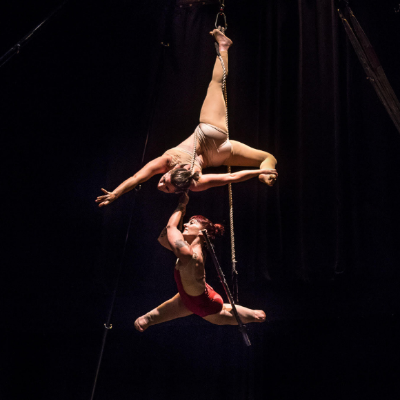 Suspended in the air, wrapped in the ropes above a trapeze bar that hangs from the ceiling, Erin, a white femme with no lower legs, splits her legs and smiles, just above the bar. Vanessa, a white woman, is upside down above Erin, hanging by one leg. The two wear outfits that match their skin and Erin’s has some red in it.