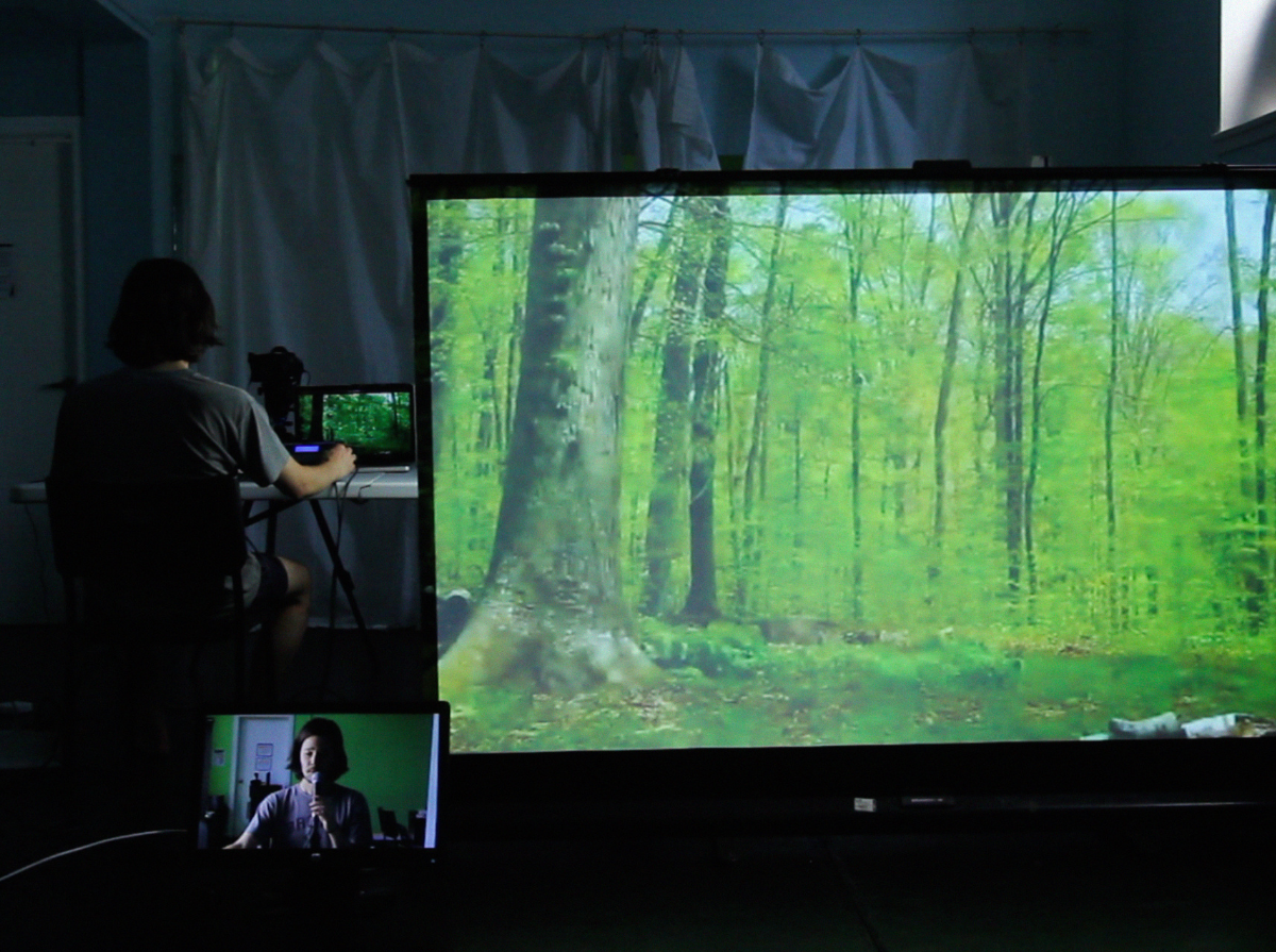 Phillip sits at a table with his back facing the camera, on the left side of a dark room. On the table, there is a computer and camera facing him. On the right side of the room, a large projector screen shows a picture of a lush green forest. In the background, there is a white door on the left side, a white drop sheet in the middle, and a production spotlight on the right side. In the foreground, a monitor shows Phillip in front of a green screen with a white door and various pieces of equipment behind him.