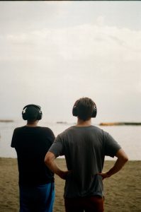 Two people wearing large headphones stand with their back to the camera facing a body of water. They stand on the sand looking over the water. The right person is wearing a grey t-shirt and stands with hands on hips. The other is wearing a black t-shirt and blue shorts with their arms down.