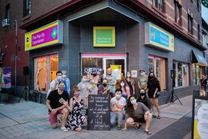A large group of people gather outside of the pop up shop. They are gathered around a chalkboard sign that reads" “ The message of what blindness knows and of what could be possible of us, of blind perception”. behind them is the pop up shop, a grey storefront space. Three signs on the storefront reading "The Pop-Up Experience" are visible. The left sign is pink, the middle sign is green and the right sign is blue.