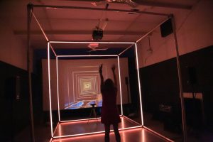 Photo of an audience member experiencing PORTAL. The image is saturated in red lighting, and the participant is standing in a large room, facing to the left with their arms over head. They are standing in a cube of yellow lighting and face a large screen with a graphic of squares.