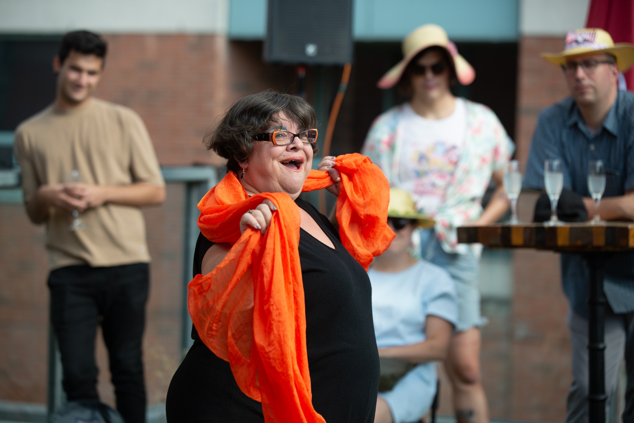 A smiling person with glasses performs wrapped in an orange scarf. Four audience members can be seen in the background behind them.