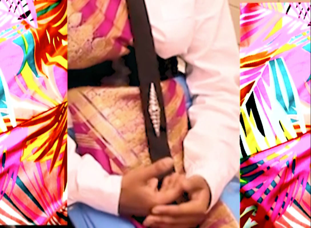 Person wearing turquoise sari over a white collared shirt, with black skinny tie with diamond shaped tie pin, brown hands folded hands in lap on either side are panels of neon palm tree print fabric.