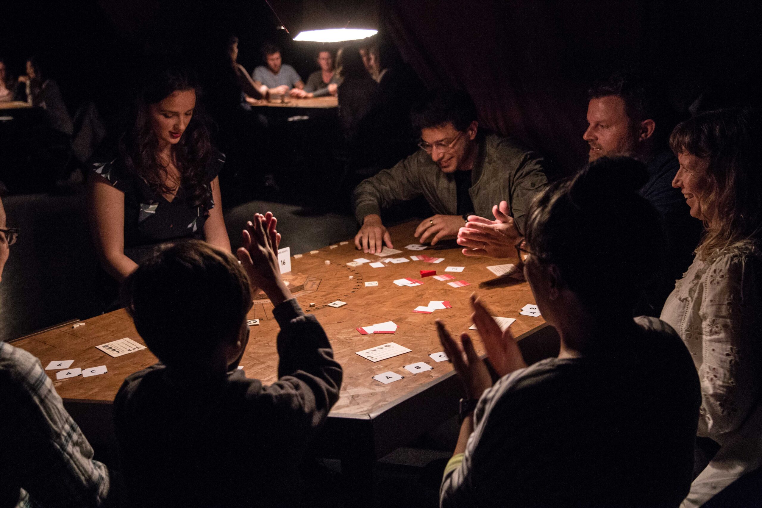 A performer smiling while looking down at a wooden game table reaches for something. Six audience members are smiling and clapping their hands in celebration of the action.