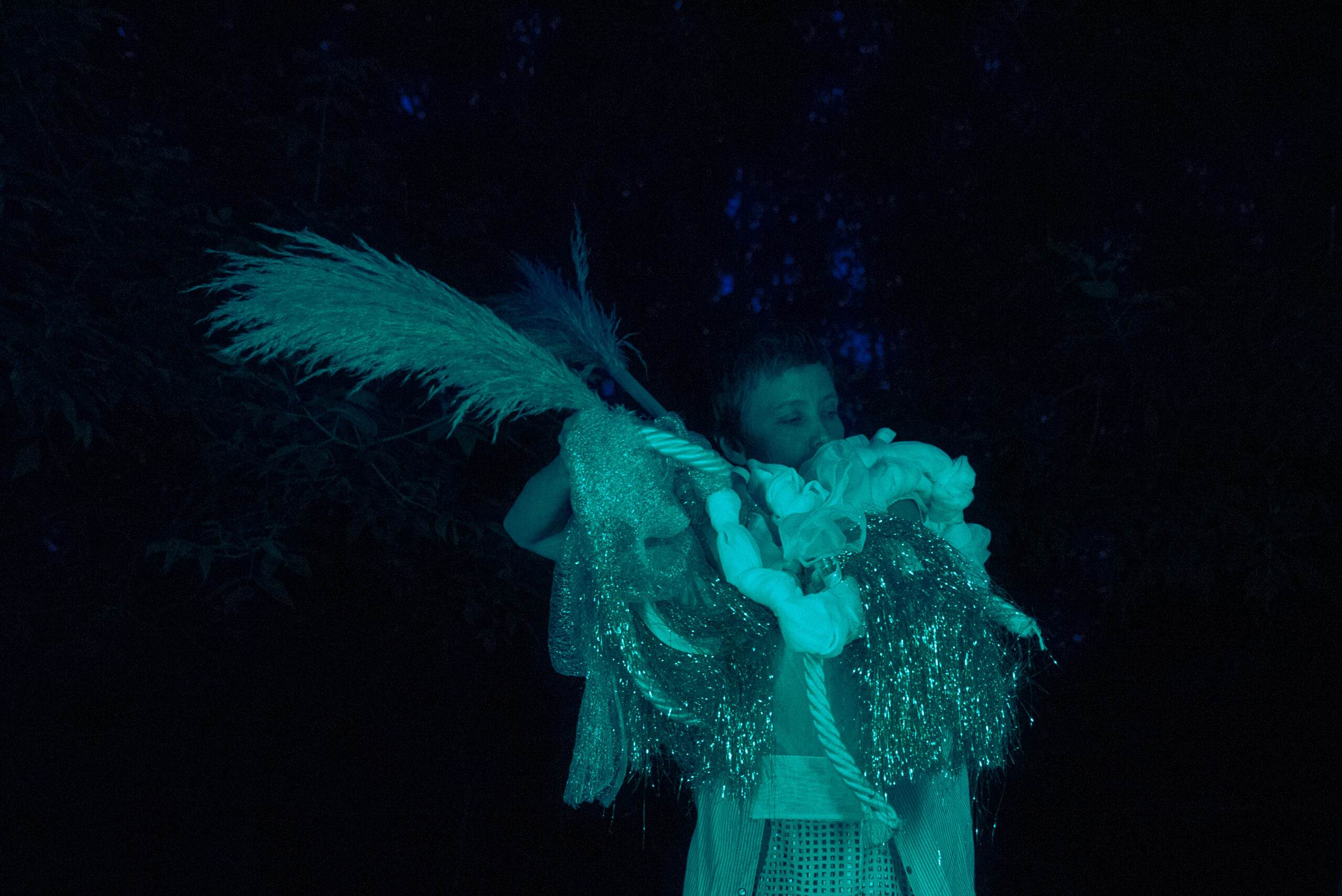 A person is holding a sculpture made from dried plants, sticks, and fabric. It is night time and the figure is lit up with green and blue light.