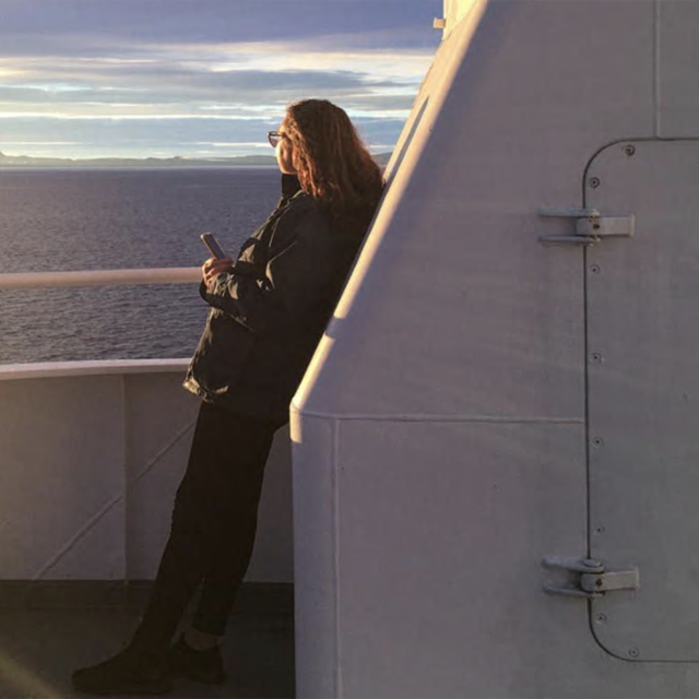 Photo of Sue on a ferry, looking out at the ocean. The sky is blue and purple, filled with wispy clouds. Sue is leaning on the ship, wearing dark clothing. She has long brown hair and is wearing glasses.