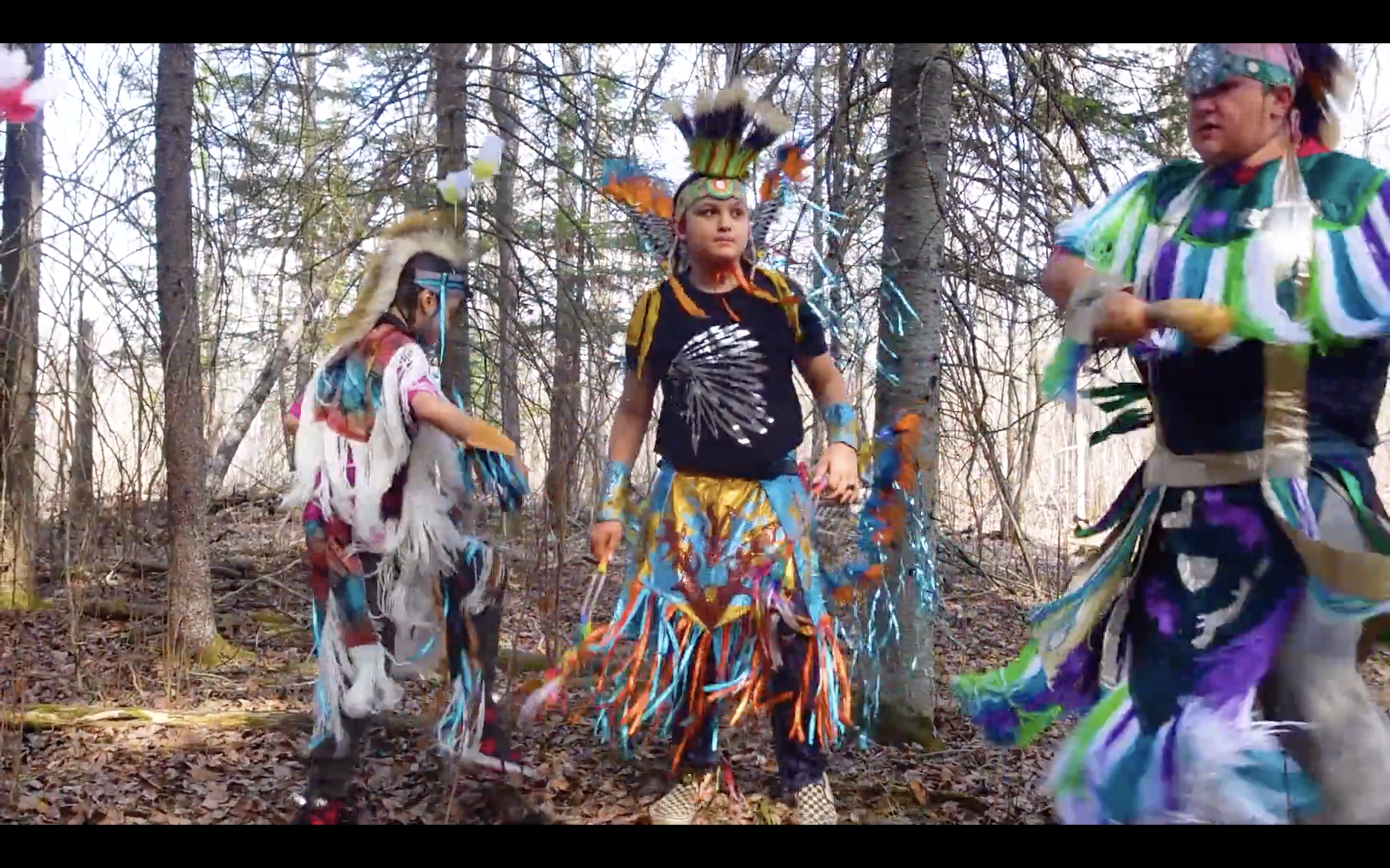 This is an image of three young Indigenous boys. They are all dressed in regalia and dancing in a forest. The boys on the left and right are dressed in colourful grass dance regalia, and the boy in the centre is dressed in fancy dance regalia.