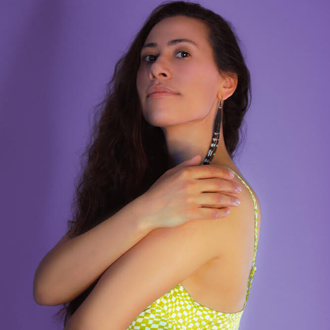 Photo of Aria, who has long, dark brown hair. They are wearing a bright yellow tank top posing in front of a lilac purple background. Their arms are wrapped around themselves, and they are looking at the camera over one shoulder.