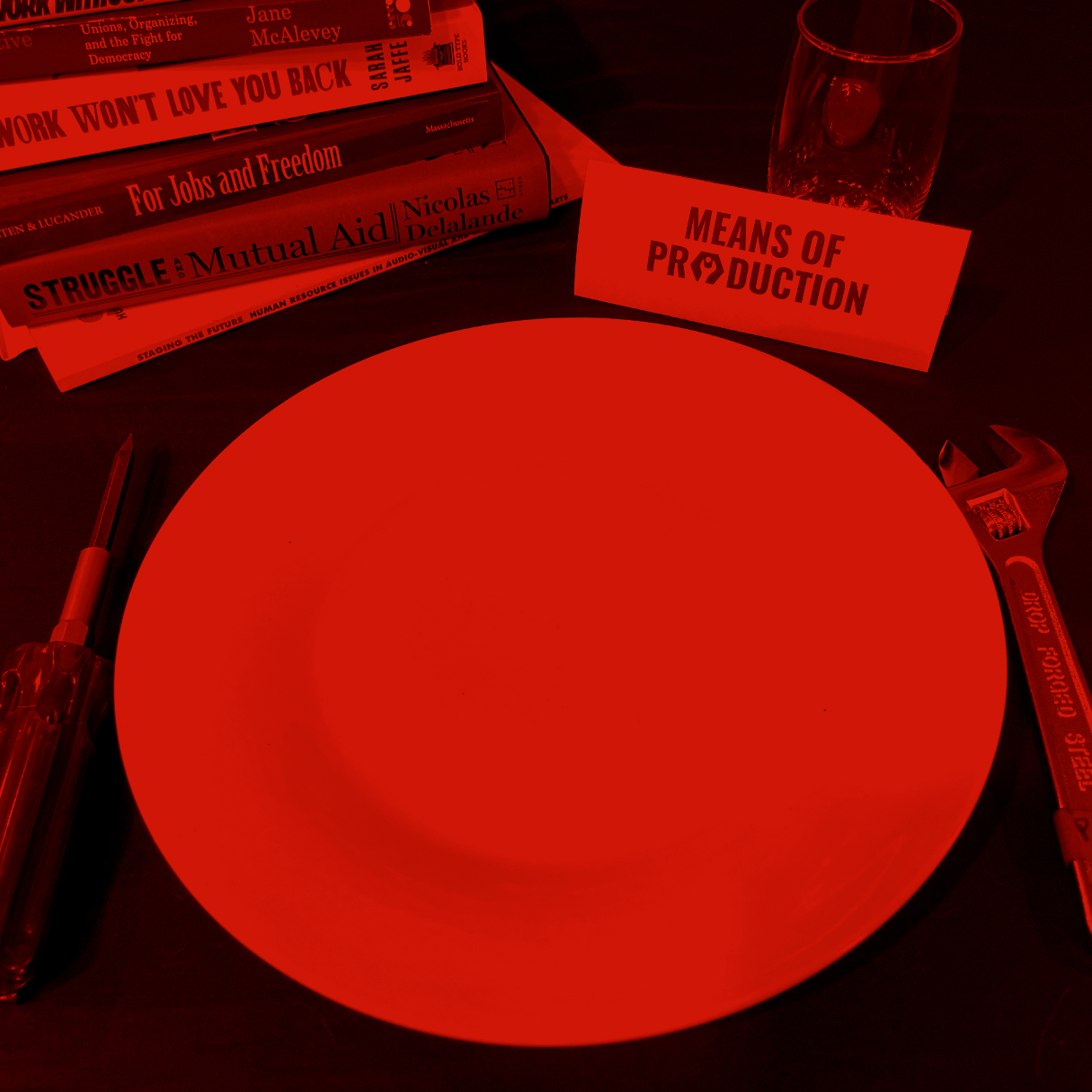A place setting is laid out on a black tablecloth. The setting includes an empty white plate, tools in the place of silverware, a stack of books, a water glass, and a placecard labelled 