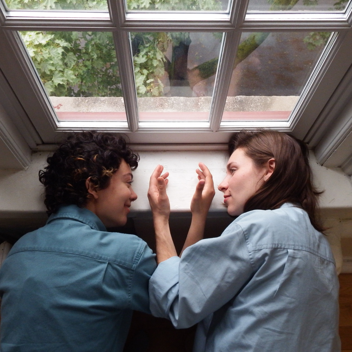Two dancers are captured by camera from above. They lay facing each other with their heads resting on a white windowsill. The dancers are softly smiling, holding their hands out with the backs of their palms in front of each other’s noses. Both dancers are wearing collared teal shirts, one lighter and one darker. The dancer on the left has short, curly dark brown hair; the dancer on the right has shoulder length straight brown hair.
