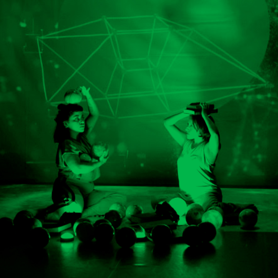 Two people sit on the ground surrounded by multiple spheres. One person cradles two of the spheres in their arm and one on their head. The other person holds two books on their head. A kaleidoscopic projection in the background with a strange geometric shape.