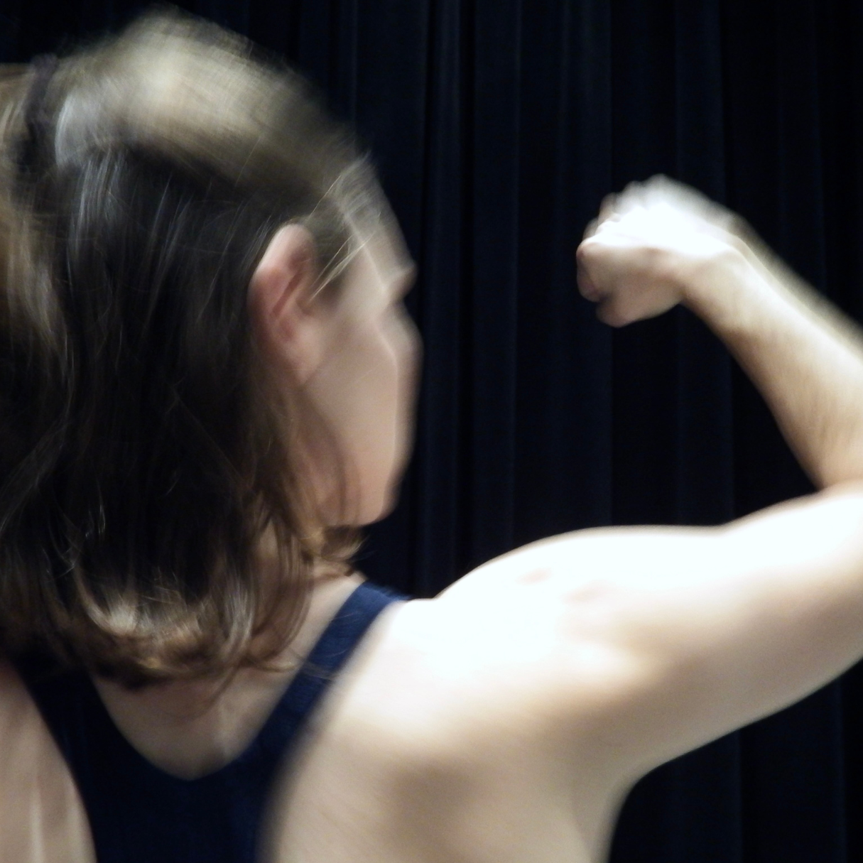 A dancer with short brown hair is captured in motion from the shoulders up in front of a black background. She is facing away from the camera and has her right first raised to eye level at her side.