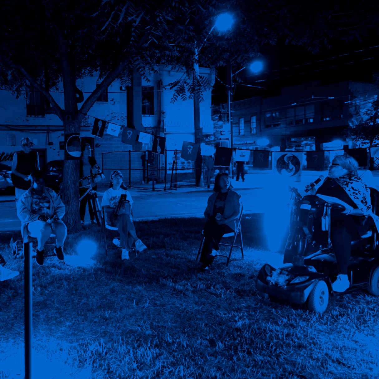 Five performers in a street-side park sit in semi-circle, lit by a purple glow. One of them is in a motorized chair and speaking into a microphone. A line of flags hangs behind them.