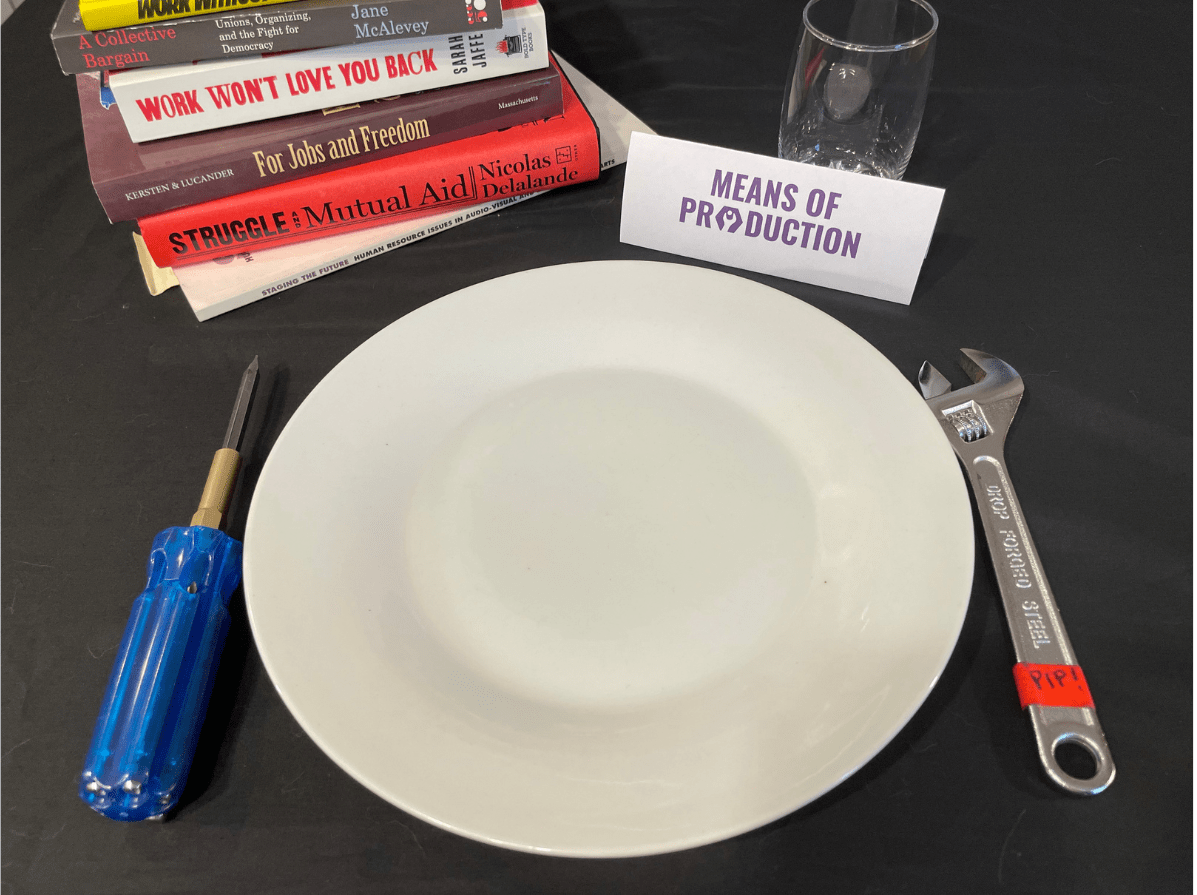 A place setting is laid out on a black tablecloth. The setting includes an empty white plate, tools in the place of silverware, a stack of books, a water glass, and a placecard labelled 