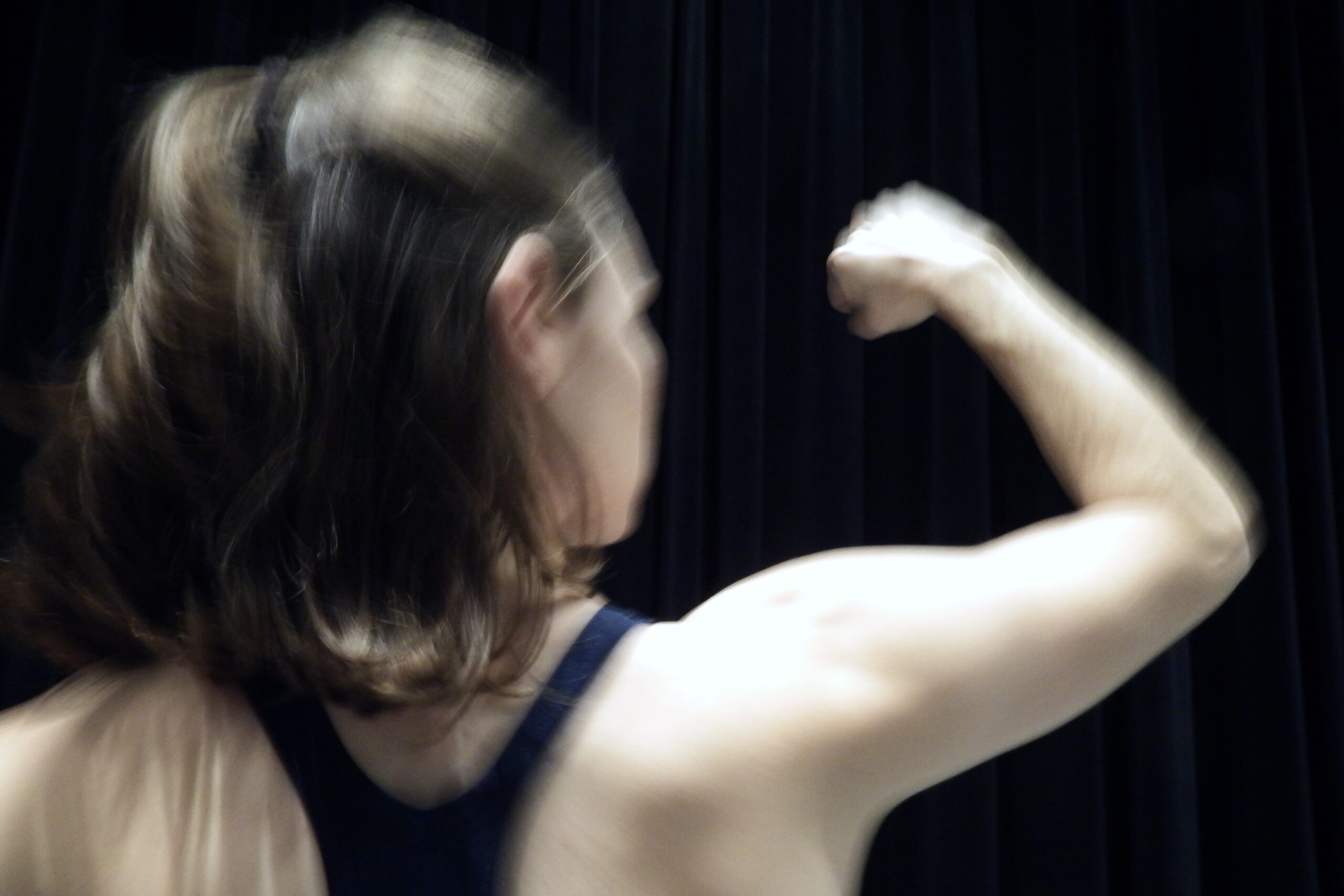A dancer with short brown hair is captured in motion from the shoulders up in front of a black background. She is facing away from the camera and has her right first raised to eye level at her side.