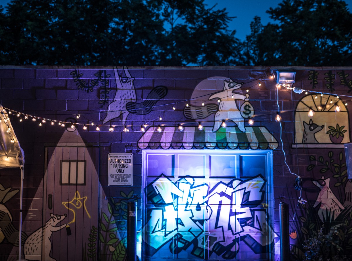 A courtyard at night decorated with strings of lights. A graffiti mural of houses and raccoons is in the background