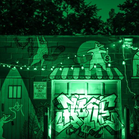 A courtyard at night decorated with strings of lights. A graffiti mural of houses and raccoons is in the background