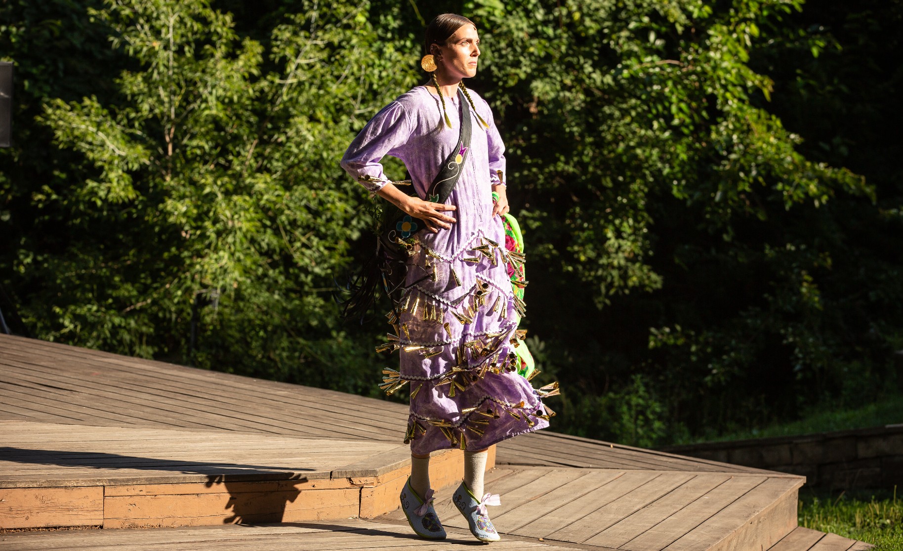 A performer stands on their toes at the edge of a wooden stage surrounded by trees. They are wearing a light purple dress with many small bells sewn onto the lower half, their long brown hair is in braids.