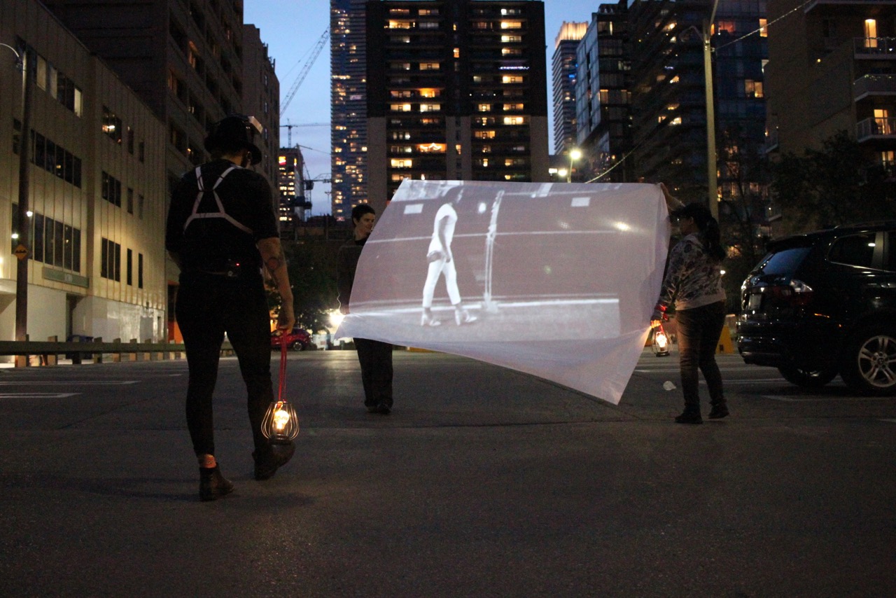 Two people are pictured holding up a white sheet with a projection of someone walking across in an urban parking lot. There is another person walking towards the projection wearing head equipment and all black, carrying a lamp in their right hand. There are different high rise buildings in the background, surrounding them.