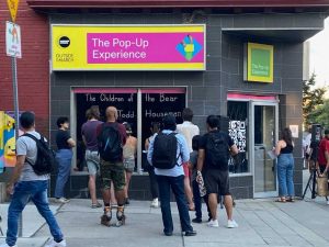 A large group of people gathered on the sidewalk outside of the storefront space. The pink and yellow sign on the storefront reads "the pop up experience" . there is writing on the window that reads "the children of the bear". The people are looking into the windows to see what is going on inside the space.