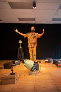 Performer stands facing the camera with their arms stretched out to the sides. They are wearing brown pants and a brown button up with a few plaid patches. behind them is a black wall. Around them are books and lamps scattered across the floor