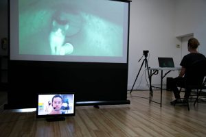 An indoor performance shot of PROOF OF EXISTENCE by Philip Nozuka. A projector is in the centre of the frame, which is displaying an image on a screen of two hands holding a blue piece of plastic. At the back, Philip sits at a desk facing away from the camera and with a laptop open in front of him.