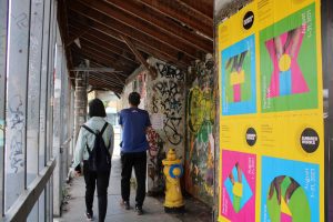 An underpass with two people walking away from the camera. Both wearing jeans, the left person has a light jean jacket, and the right person has a blue t-shirt. They walk away from the camera past a yellow fire hydrant. In the right of the image four summerworks posters are hanging on the wall.