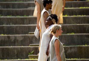 An outdoor performance shot of nowhen by Alison Wong. Several performers are standing in a line that goes up the steps of the High Park Amphitheatre. They are staggered to be facing out to the left or right, all are wearing loose-fitting light fabrics and have their arms at their sides.