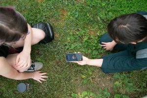 two people are crouched down over grass, with the photo taken from above them looking down. The person on the right is wearing a green jumper, and holds a phone out in her hand between them. They are listening to something on the phone. Both people have dark brown hair.