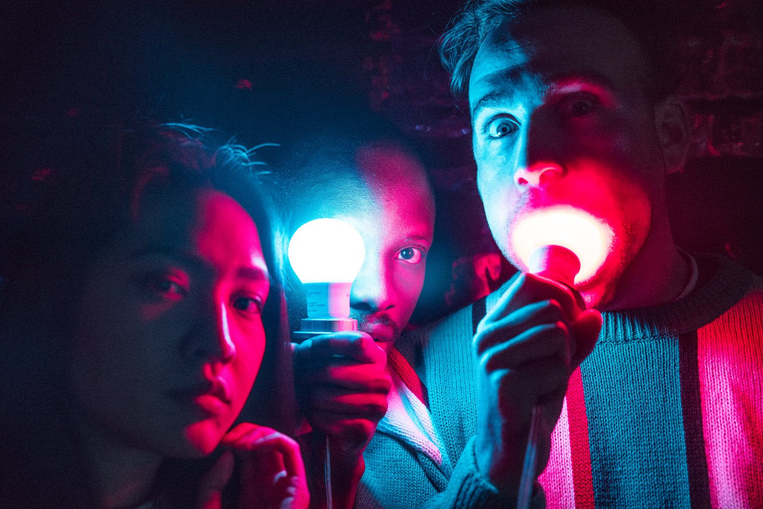 three people in shades of red and blue hold two lightbulbs near their faces