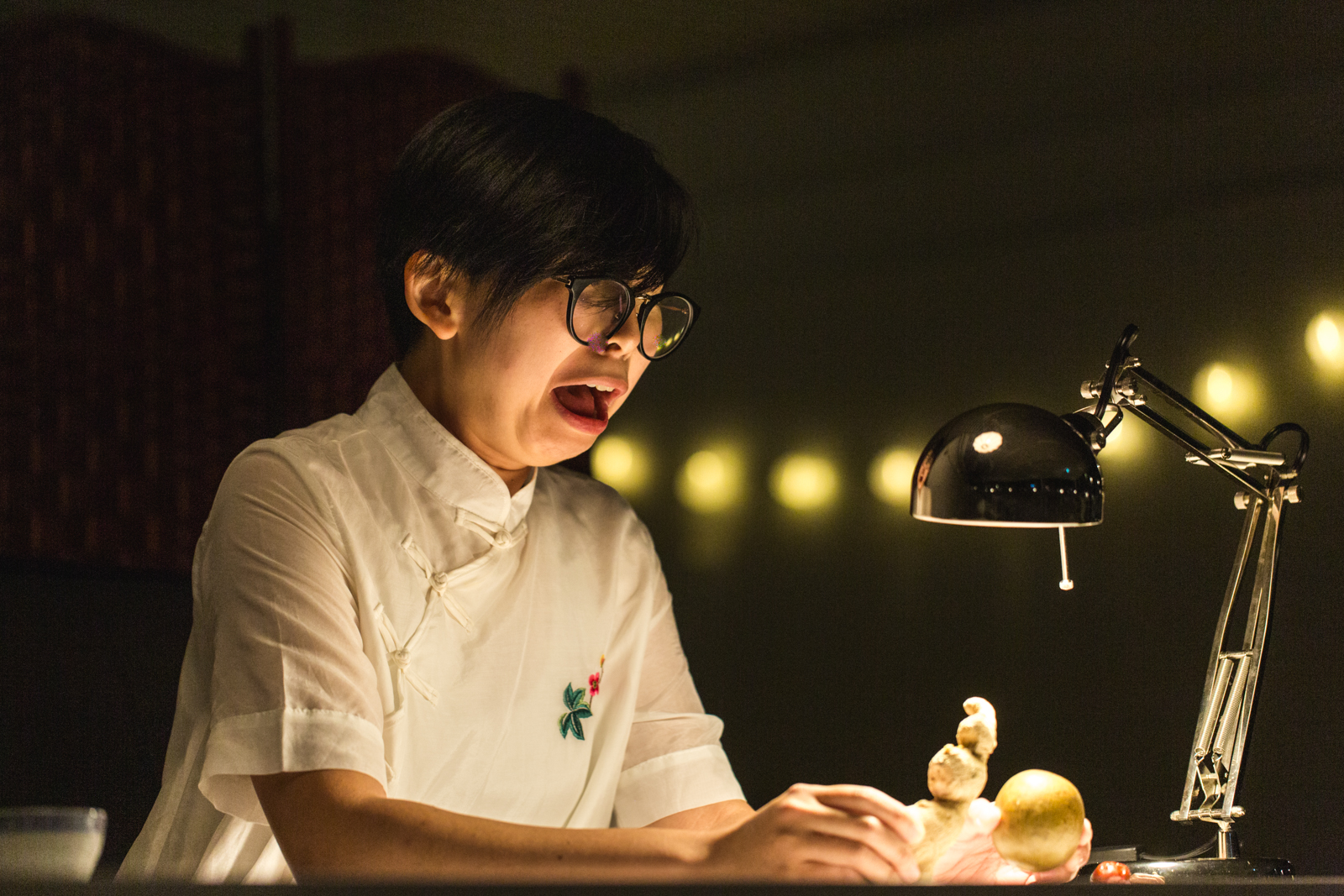 Gloria Mok has short black hair, black rimmed glasses, and is wearing a white dress with a pink and green embroidered flower on her chest. She is holding a ginger root and a monk fruit as if they are puppets. The background is black with a string of warm glowing lights in the distance.