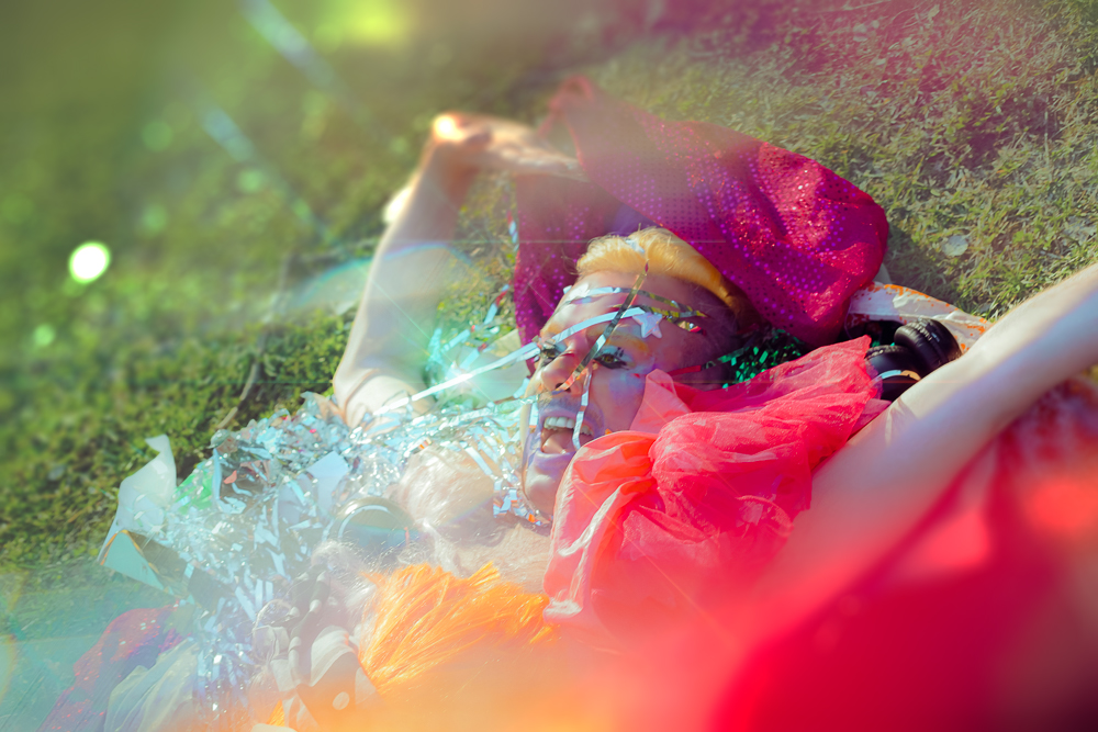 STARLIGHT is laying in the grass in a pile of brightly-colored fabric and shiny tinsel. He has brightly coloured smudged makeup on his face and is smiling wide with delight. The photo is slightly blurred giving motion