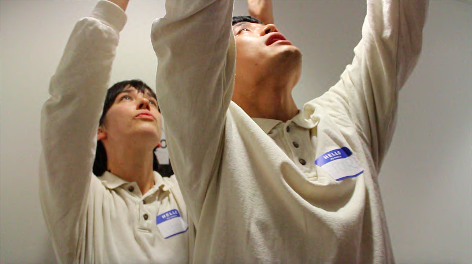 This is a medium wide photo of two people wearing ugly beige button up polos buttoned all the way to the top.They are standing with Roberto a… (description here) in the foreground and Devon a….(description here) in the background in a room that looks like a convention centre. There is a projection screen, round tables, chairs, speakers and a nondescript black curtain that are all slightly out of focus. The two performers are both standing making the same pose, hands clasped in front of their bodies with expressions that look as though they are going to announce a delightful detail about the space they are in.