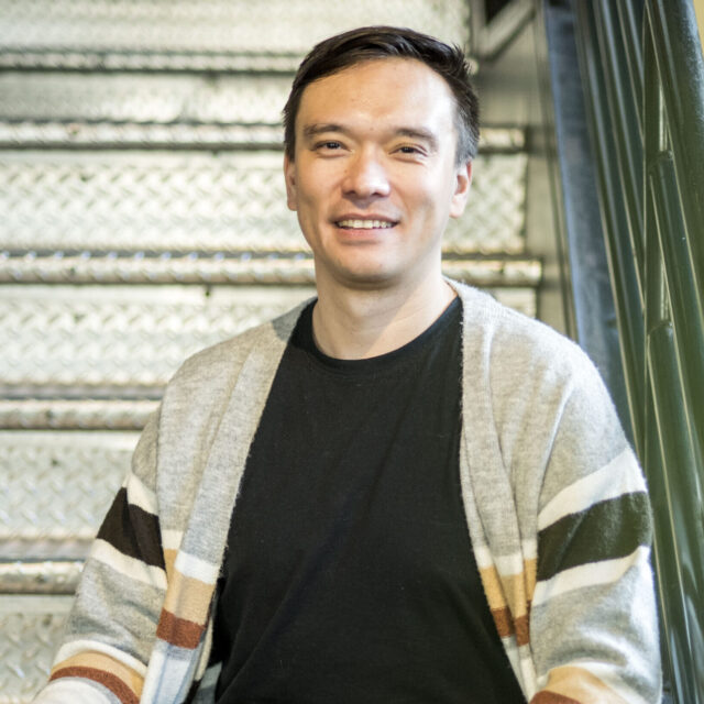 Image of Michael Caldwell, an Asian man with dark brown hair wearing a black t-shirt and a grey cardigan with green, cream, and dark orange stripes on the sleeves. He is sitting on a metal staircase that has a green handrail. He is looking directly at the camera, smiling.