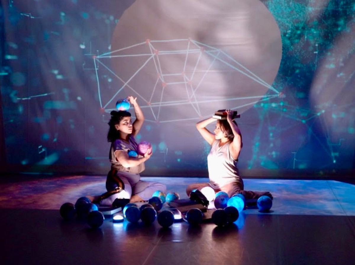 Two people sit on the ground surrounded by multiple spheres. One person cradles two of the spheres in their arm and one on their head. The other person holds two books on their head. A kaleidoscopic projection in the background with a strange geometric shape.