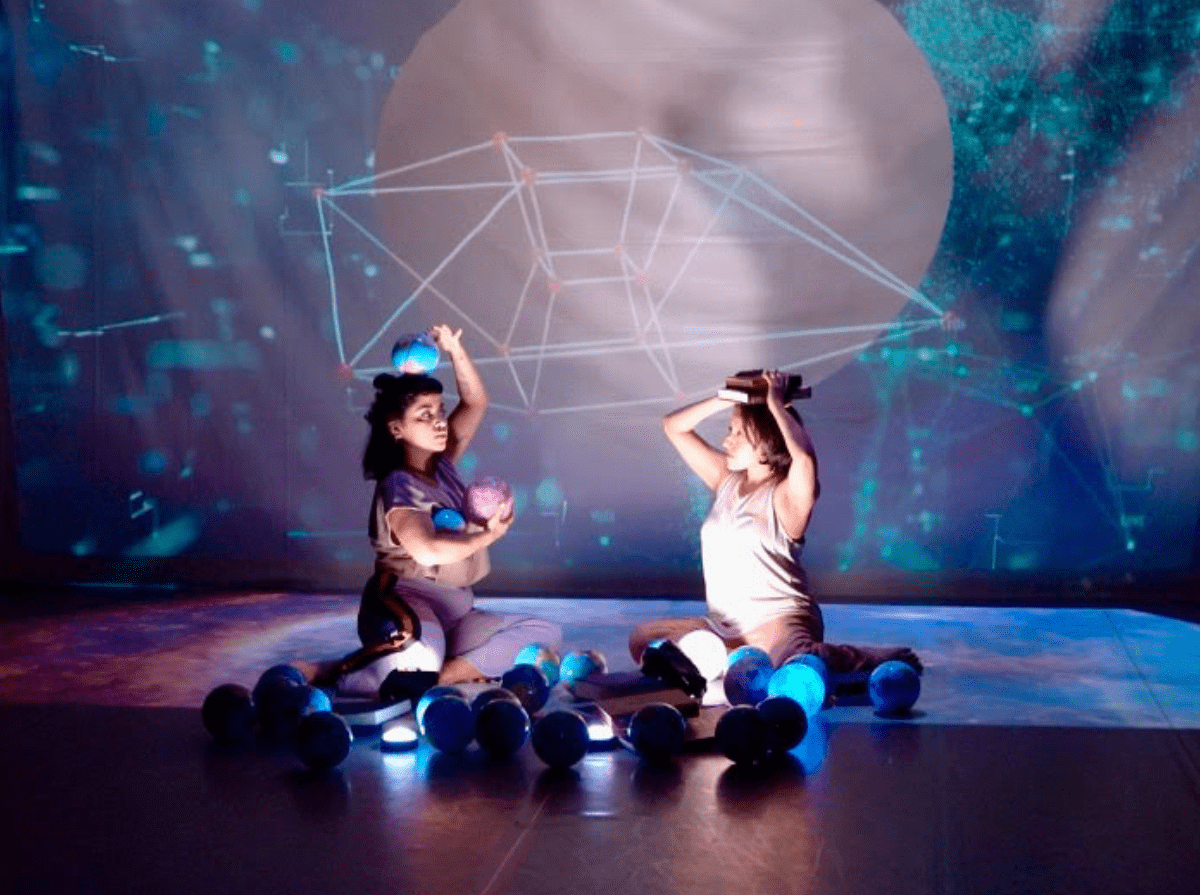 Two people sit on the ground surrounded by multiple spheres. One person cradles two of the spheres in their arm and one on their head. The other person holds two books on their head. A kaleidoscopic projection in the background with a strange geometric shape.