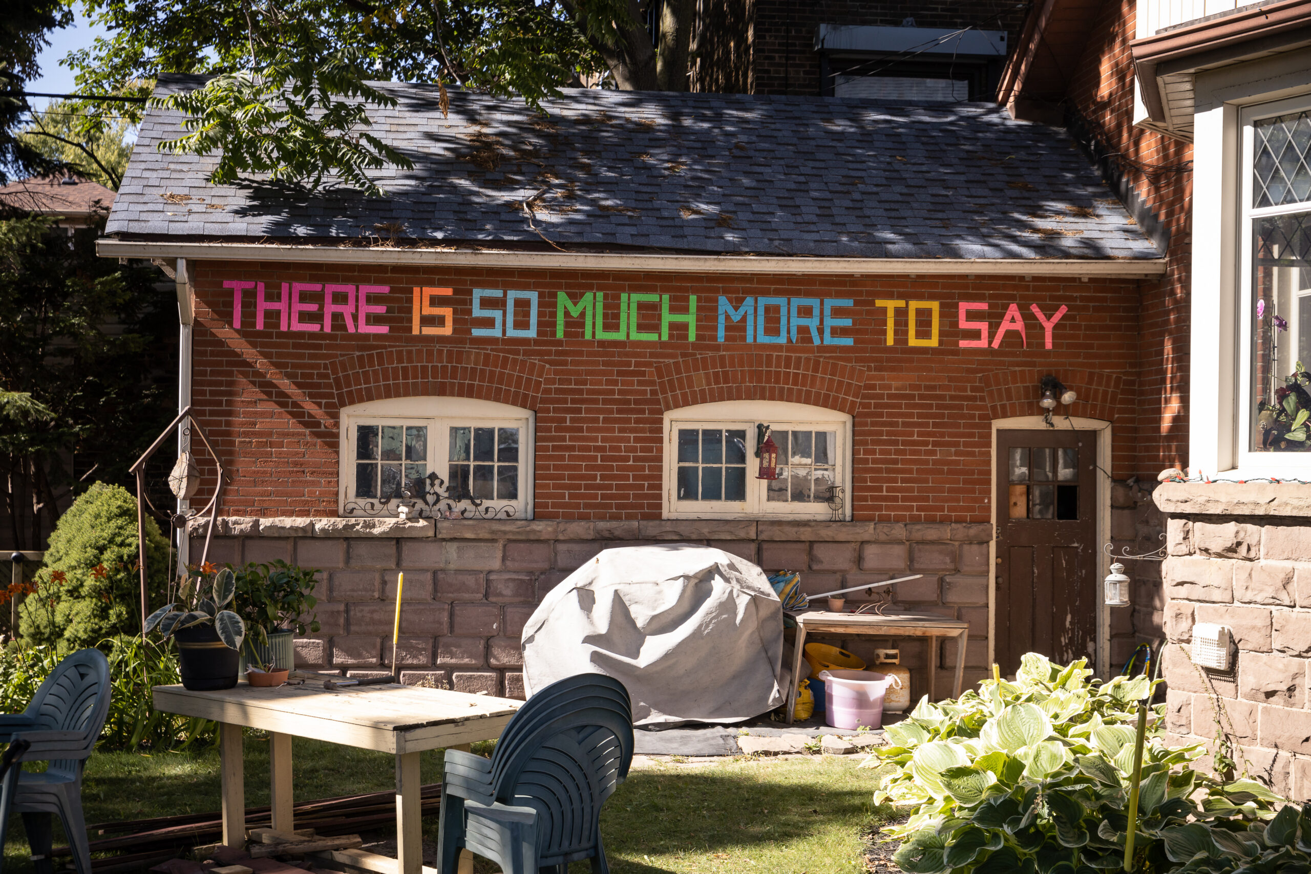 The words 'There is so much more to say' is written in colourful tape on the brick wall of a residence in the daytime.