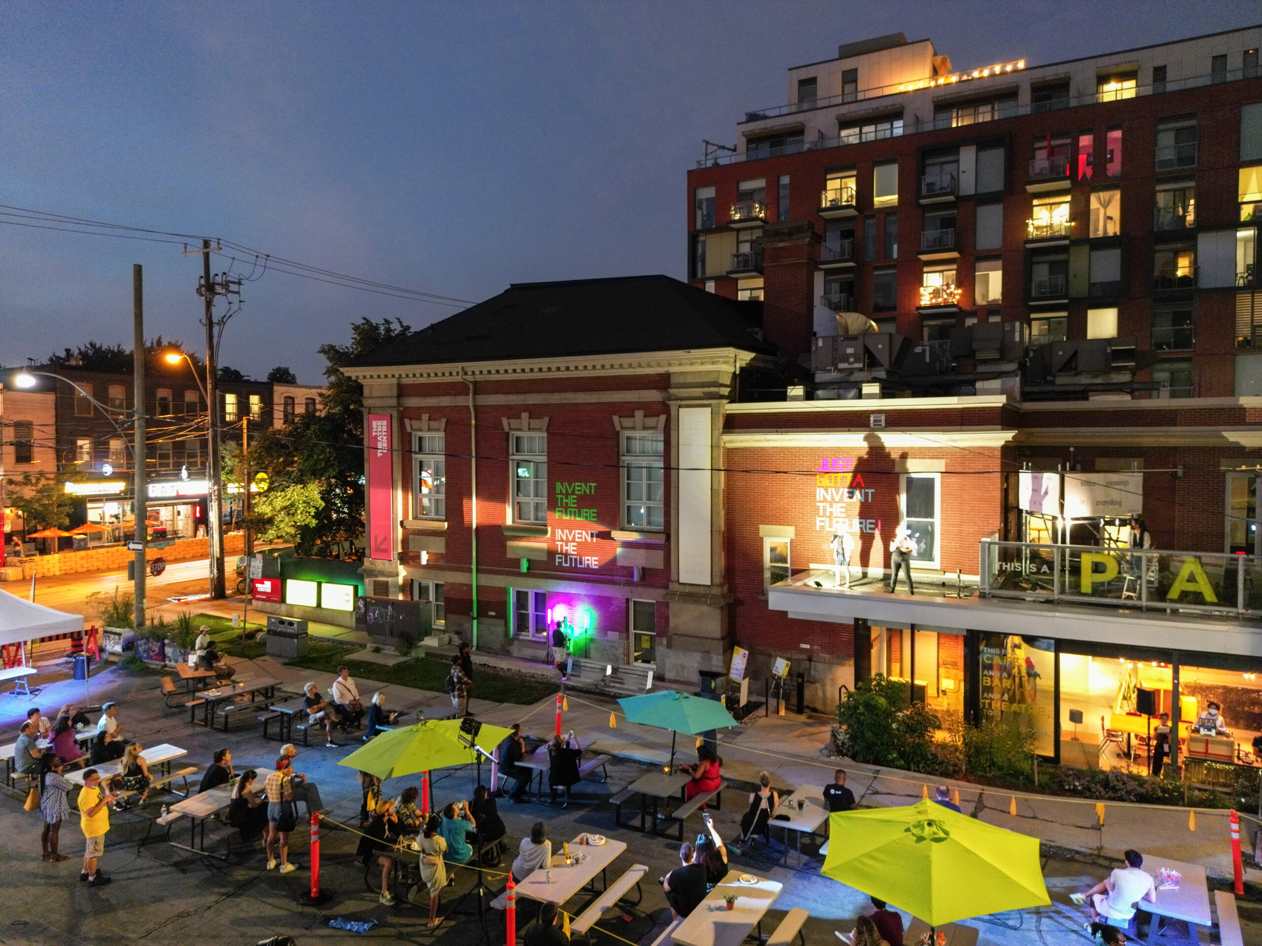 A theatre lit up at night with the words 'Invent the Future' on the wall and a spotlight on a speaker standing on a balcony. On the ground, spectators look up from picnic tables and neon green umbrellas.