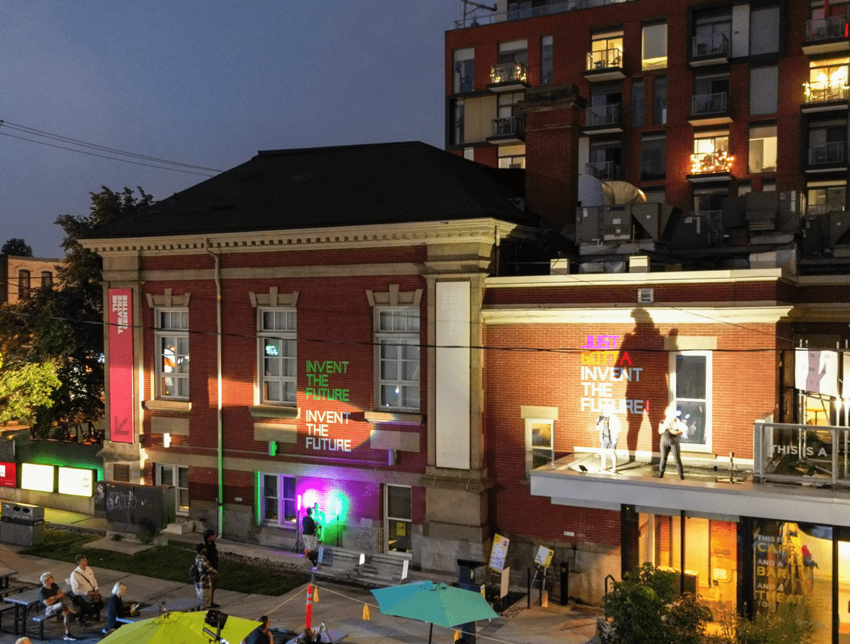 A theatre lit up at night with the words 'Invent the Future' on the wall and a spotlight on a speaker standing on a balcony. On the ground, spectators look up from picnic tables and neon green umbrellas.