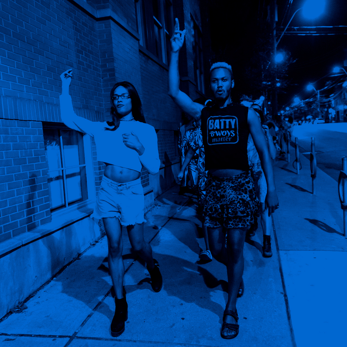 Two black performers with their fists raised are leading the audience down a sidewalk. One has short orange hair, a cheetah print skirt and a black shirt saying 'Batty Bwoys Matter'. The other has long black hair and is wearing a white crop top with jean shorts.