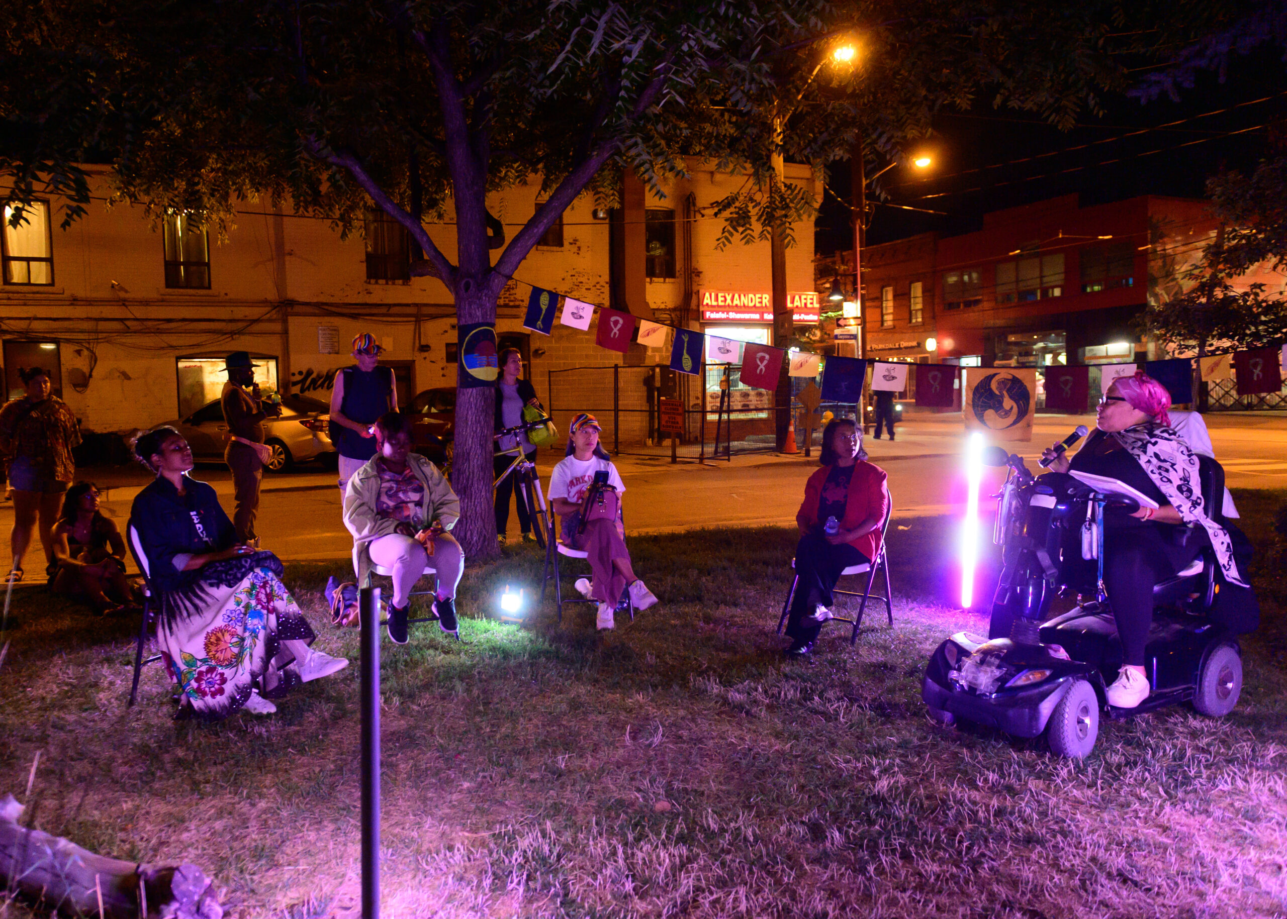Five performers in a street-side park sit in semi-circle, lit by a purple glow. One of them is in a motorized chair and speaking into a microphone. A line of flags hangs behind them.