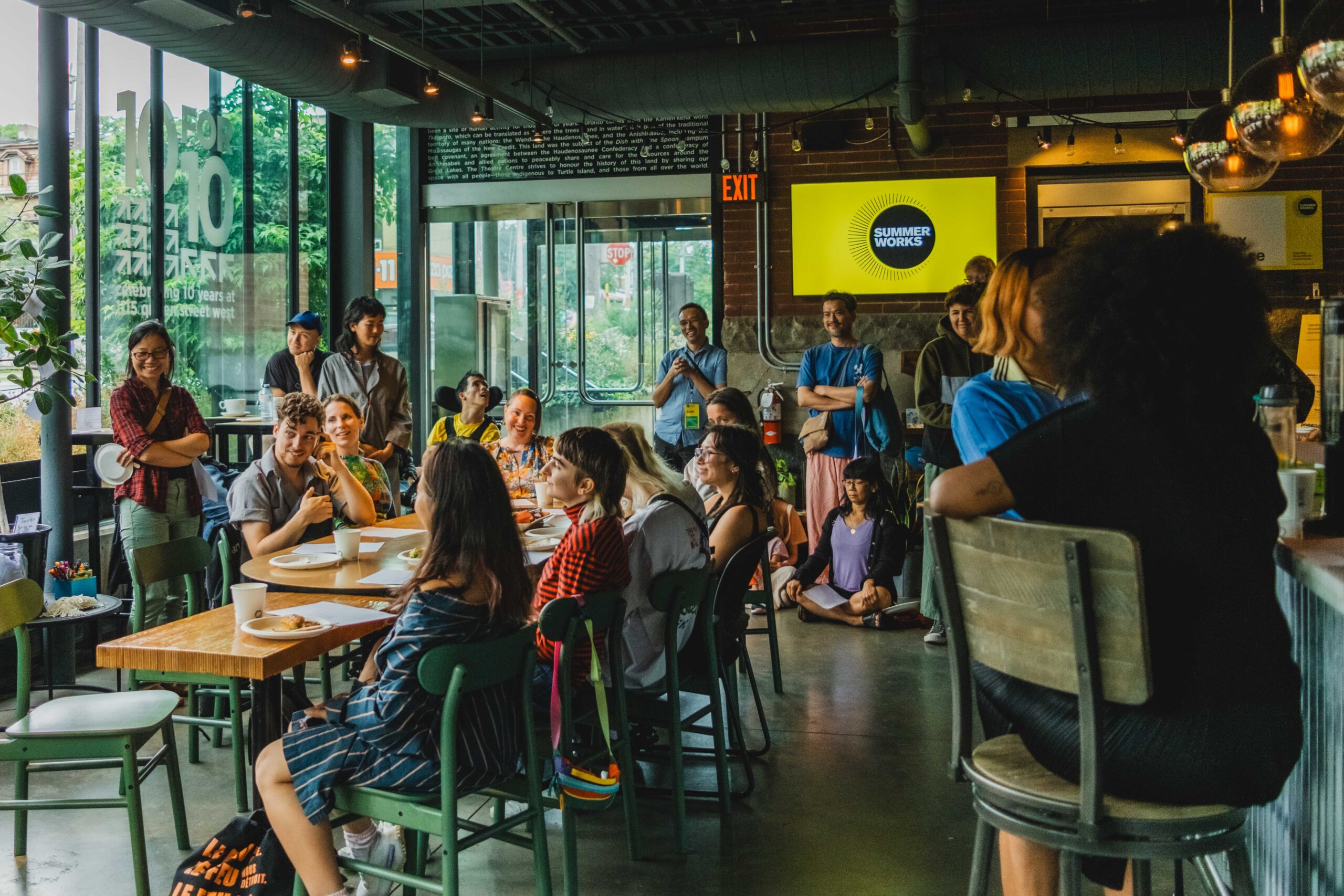 A group of people gathering in a theatre lobby, sitting and standing, with the SummerWorks logo on a TV screen in the background